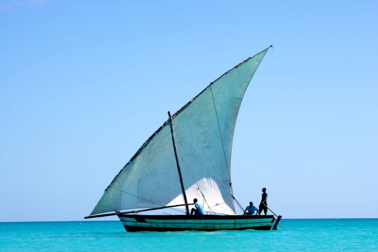 Ein Segelboot mit drei Personen im Wasser, dahinter blauer Himmel