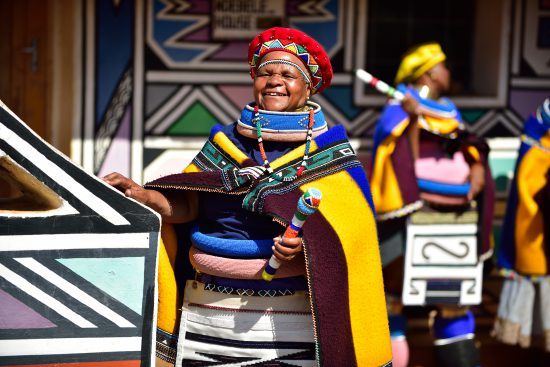 Eine Frau in bunter Ndebele-Kleidung lächelt in die Kamera, weitere Frauen sowie farbenfrohe Kunst im Hintergrund