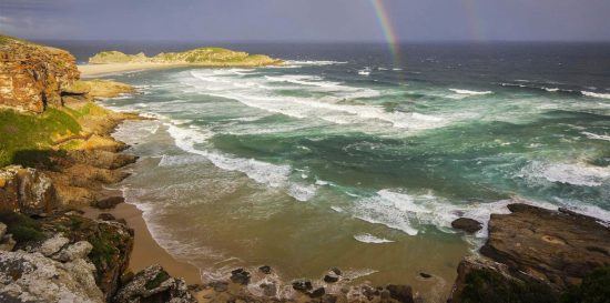 Ein Regenbogen über einem stürmischen Meer, das auf einen Sandstrand und Felsen trifft