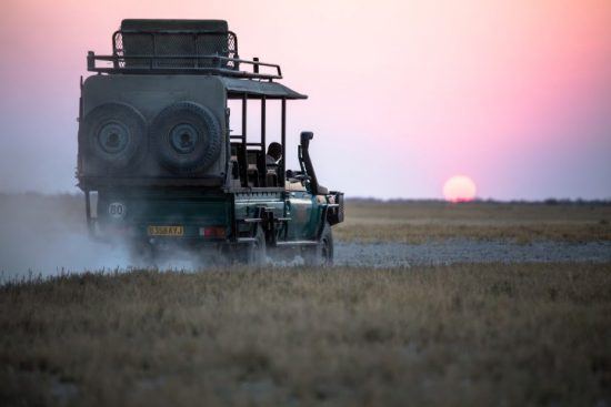 Ein vollbepackter Jeep fährt in der offenen Ebene Botswanas in einen rosa Sonnenuntergang