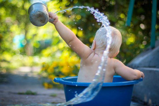 Baby badet in einer blauen Wanne und gießt Wasser im hohen Bogen aus