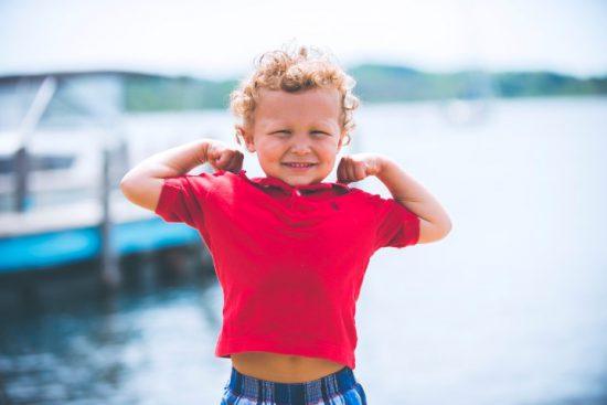 Ein Junge in einem pinken Shirt präsentiert seine Muskeln vor einem Bootsanleger