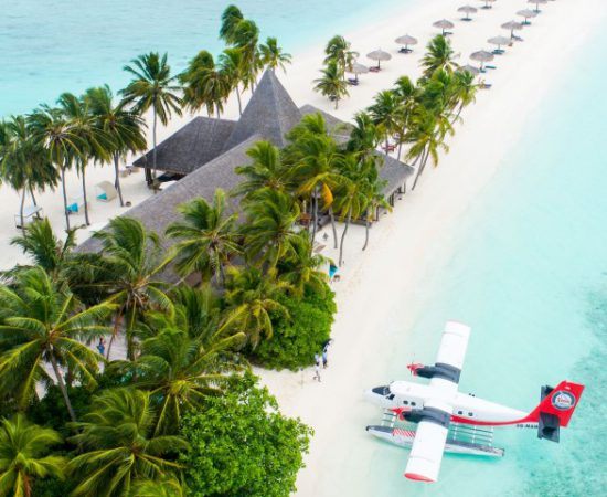 Urlaub auf den Malediven: Ein Wasserflugzeug landet an einem weißen Strand mit Palmen und einem Gebäude