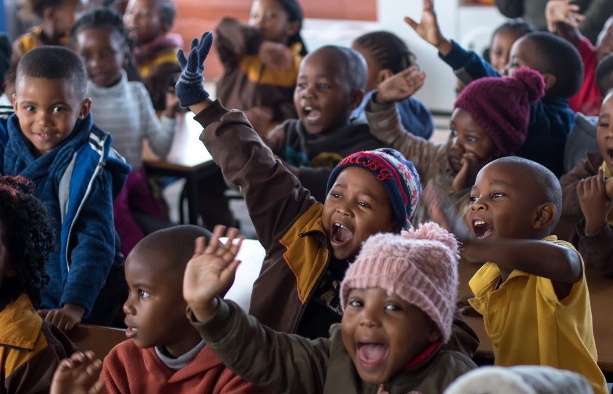 Kinder sitzen bei einer Versammlung zusammen und strecken ihre Arme in die Luft