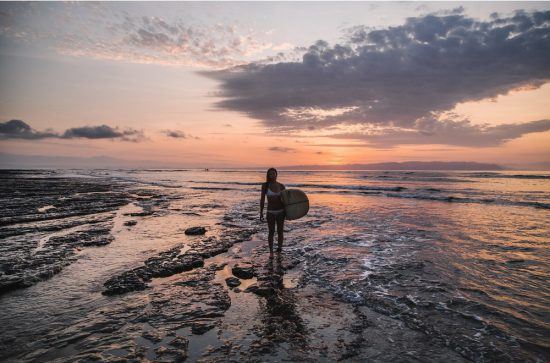 Patricia Pinto, the founder of The founder or Love Surf Yoga, Patricia Pinto Photo Credit: Love Surf Yoga on the beach with a surfboard