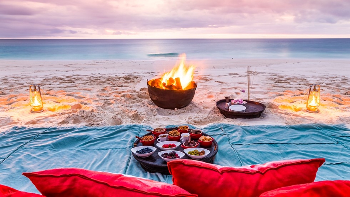 Picnic setup with a blue blanket, red pillows, tapas and a bonfire at sunset