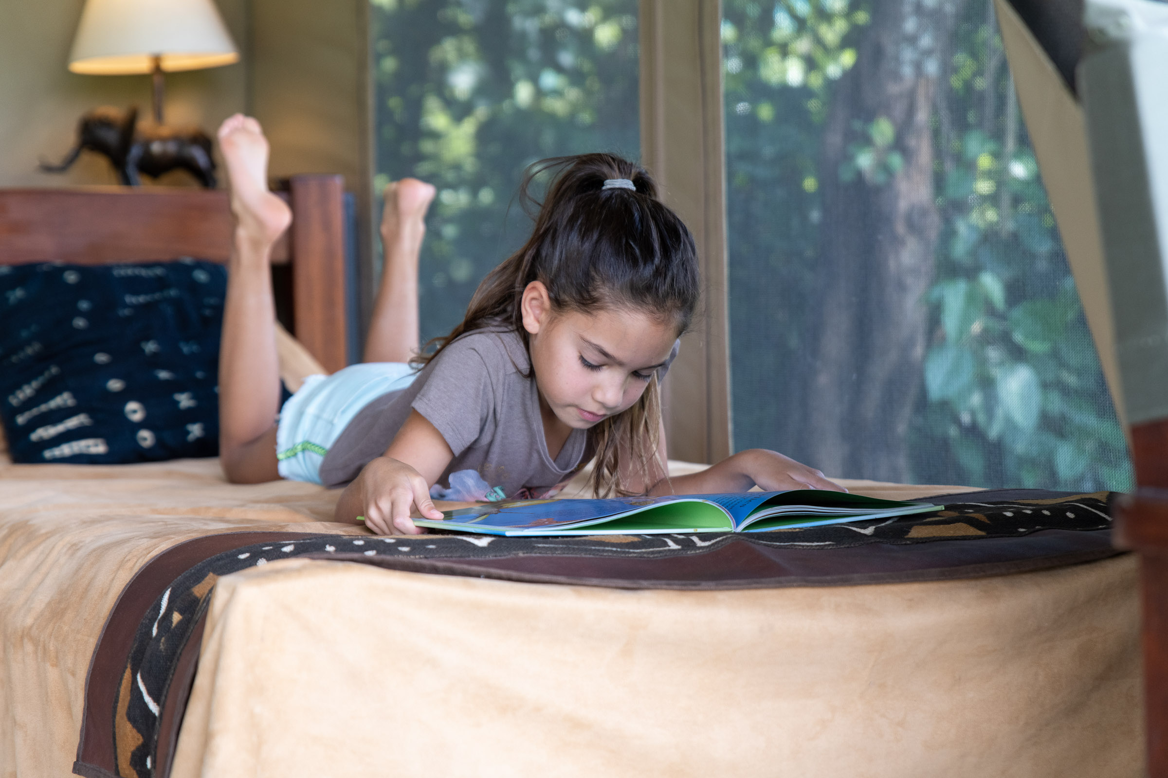 Little girl reading a book on her bed in a safari tent