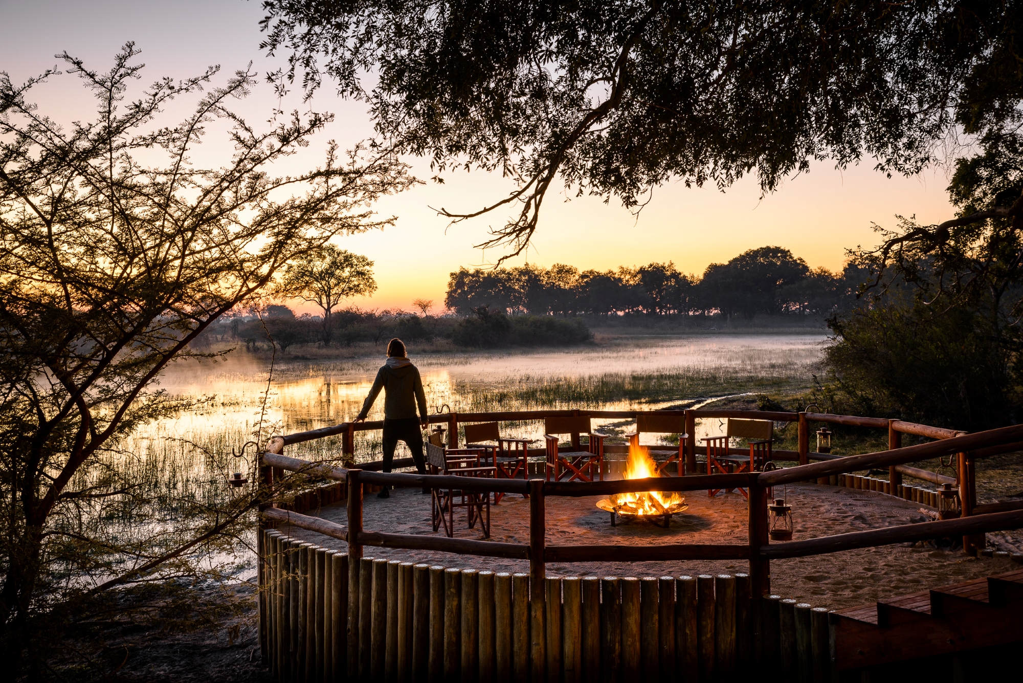 Man standing by an elevated firepit area overlooking a river at sunset